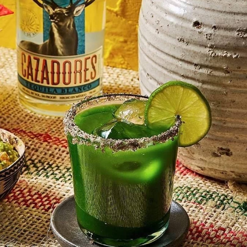Green margarita with a lime slice and salted rim, next to a tequila bottle and guacamole.