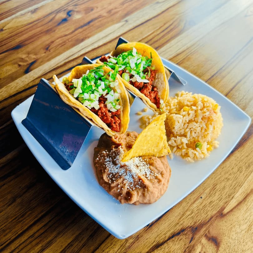 Two El Sazón tacos with rice and refried beans, garnished with cheese and a tortilla chip.