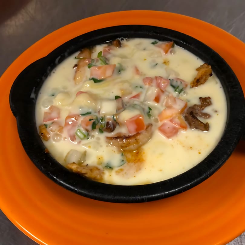Melted white cheese dip with diced tomatoes, onions, and meat in a black bowl on an orange plate.