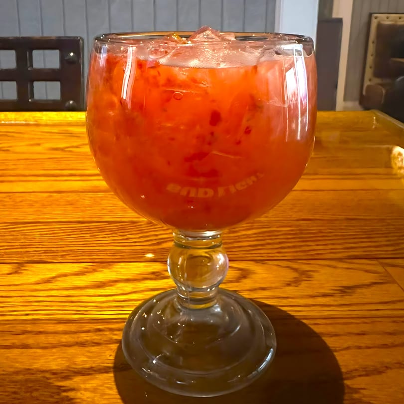 A large glass of red, fruity Mexican drink with ice sits on a wooden table.