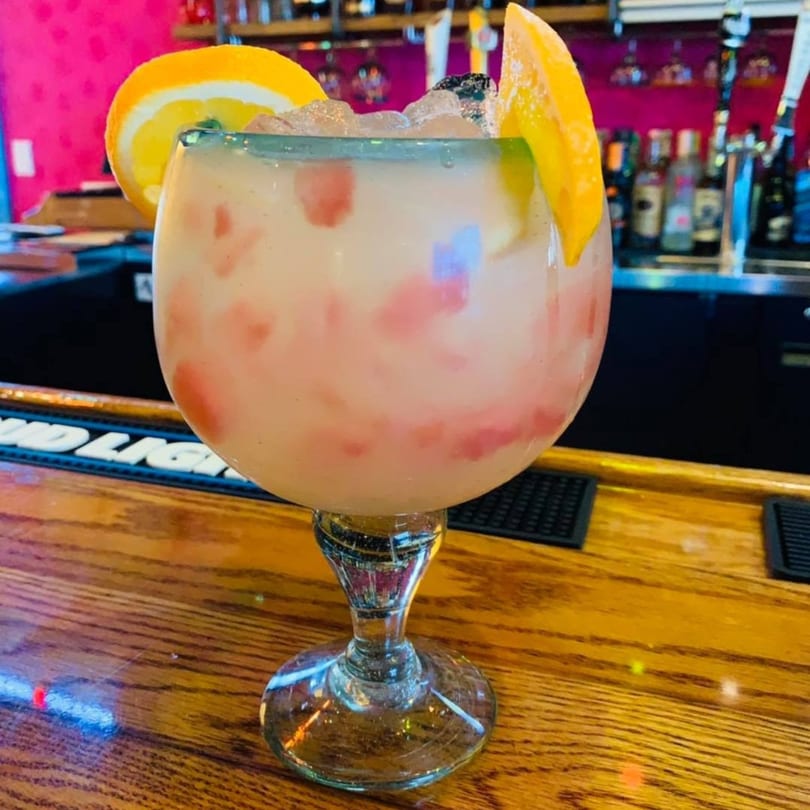 A large, light pink margarita with fruit chunks, garnished with orange slices, on a wooden bar.