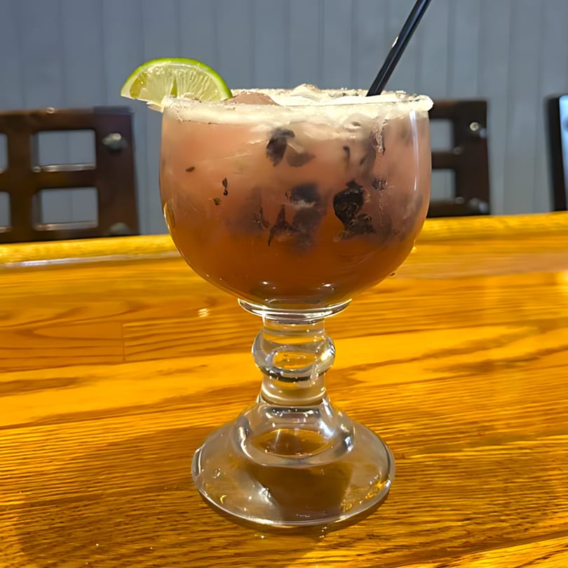 A dark red margarita with a salted rim, lime wedge, and black straw on a wooden bar.